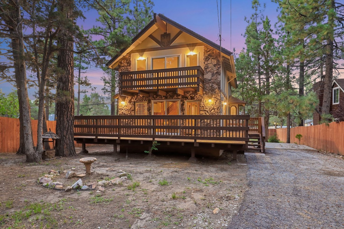A two-story cabin features a stone façade and wooden balcony railings. A spacious front deck is surrounded by tall trees, with a gravel path leading to the entrance. A fire pit made of stones is placed on the ground, enhancing the outdoor space.
