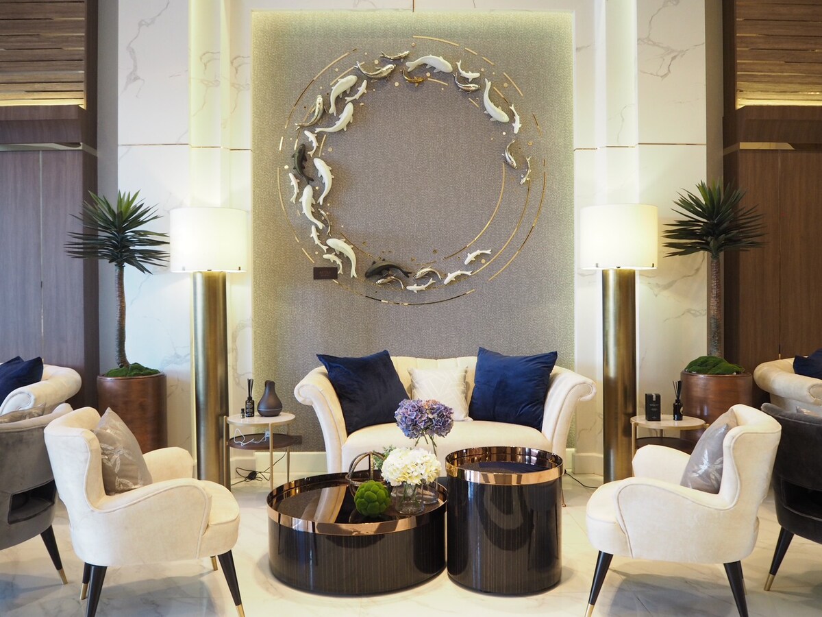A stylish seating area features two plush armchairs and a comfortable sofa placed around circular coffee tables. A decorative wall art piece with a circular design hangs prominently, complemented by modern floor lamps and potted plants on either side.