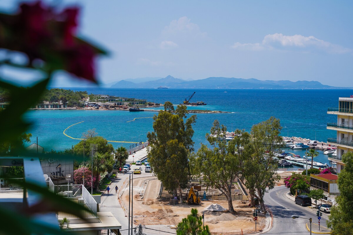A vibrant view captures the bustling marina and the sandy beach, framed by lush greenery. The bright blue sea meets the sky in the distance, while construction activity is visible along the shoreline. The image showcases a mix of natural and urban elements.