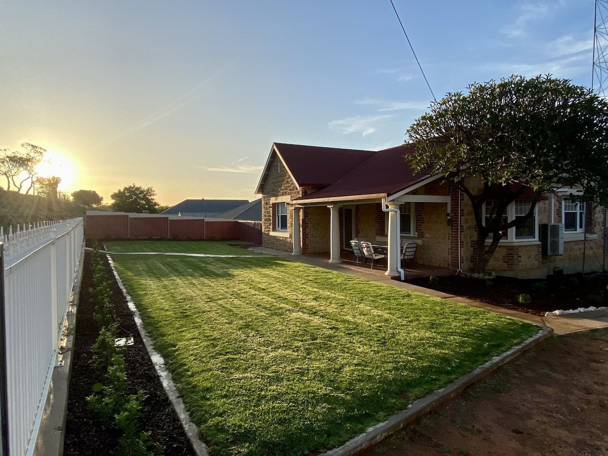 A charming stone home is set against a serene sunset. The well-maintained lawn features a smooth green expanse, bordered by flower beds. A covered porch offers seating, while a white picket fence enhances the outdoor space. Trees provide additional shade and visual appeal.