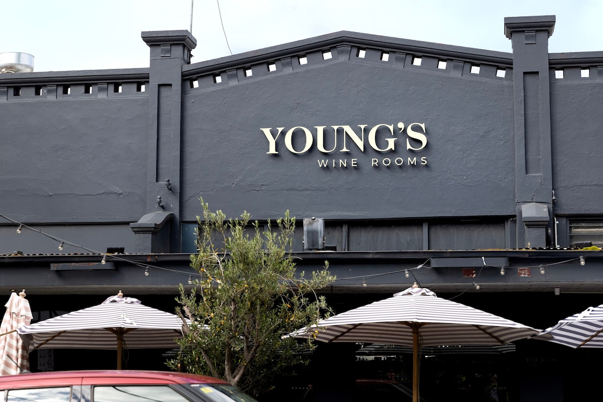 The exterior of Young's Wine Rooms is depicted, featuring a dark facade with large lettering identifying the venue. Outdoor seating is visible with striped umbrellas providing shade. Decorative plants are positioned at the entrance, offering a welcoming touch.