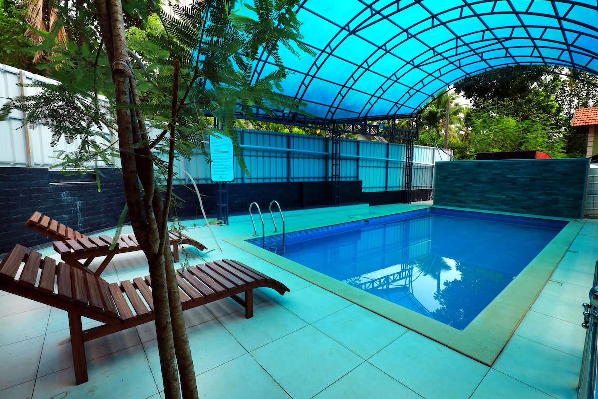 A covered swimming pool area features a clear blue roof that allows natural light. Wooden lounge chairs are placed beside the pool, offering a comfortable spot to relax. Lush greenery is visible in the background, enhancing the tranquil setting.