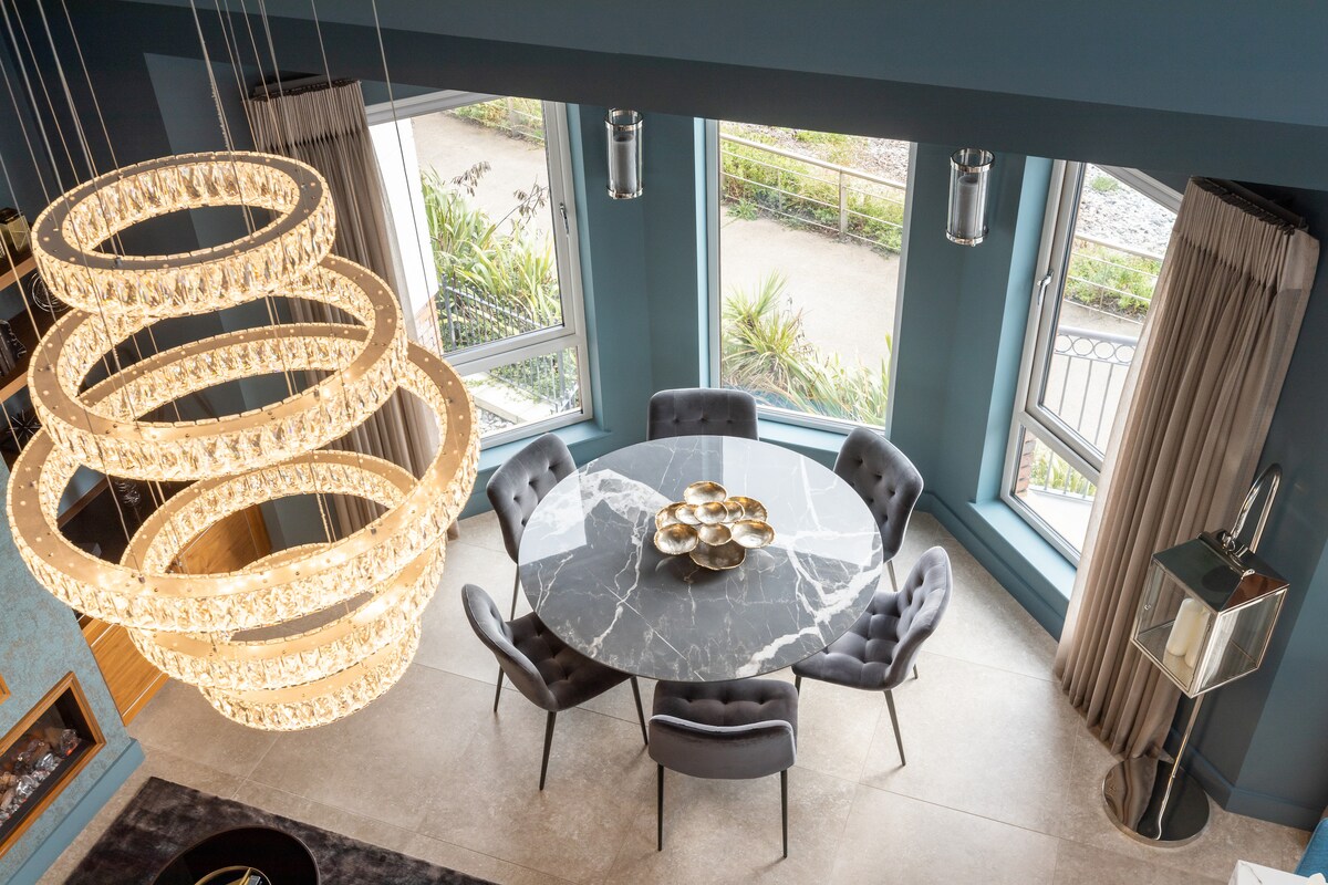 A modern dining area is highlighted by a round marble dining table surrounded by plush gray chairs. Large windows offer natural light and views of the outside greenery. A striking, artistic chandelier hangs overhead, adding elegance to the space.