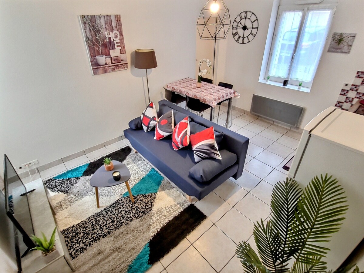 A modern living area features a spacious sofa adorned with decorative cushions, complemented by a colorful area rug. A dining table is arranged nearby, with four chairs positioned around it. Natural light fills the room through a set of windows, enhancing the inviting ambiance.