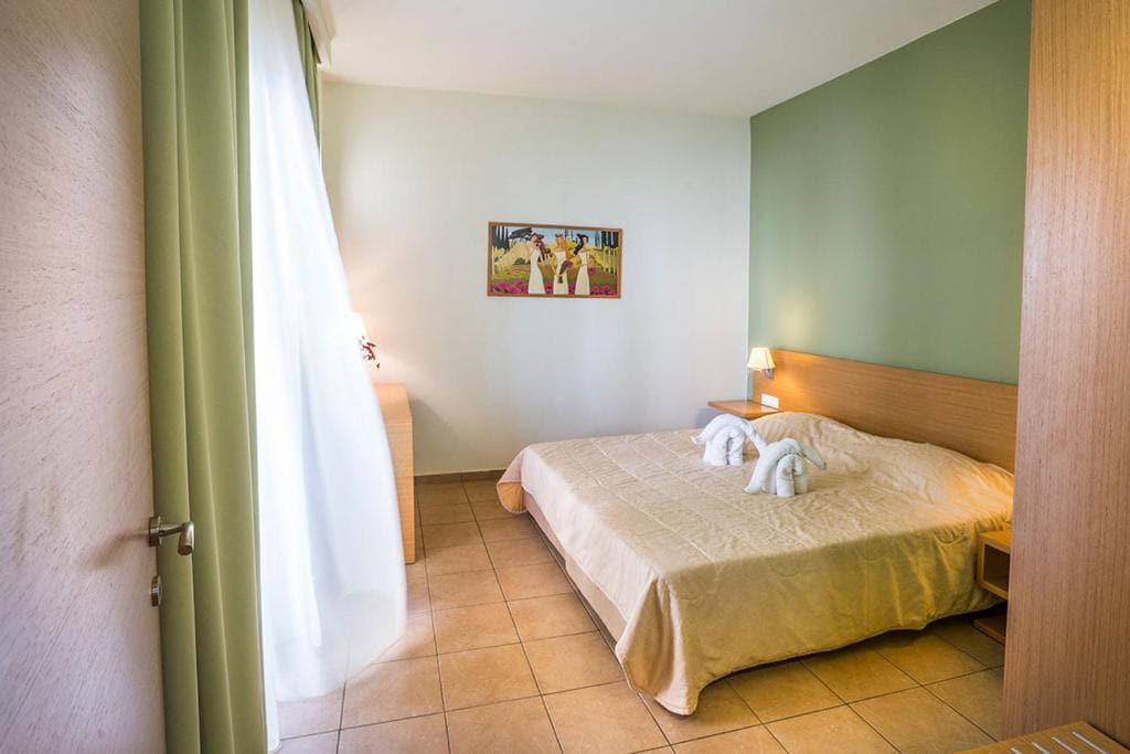 A spacious sleeping area features two single beds or a double bed, covered in a soft beige blanket. Natural light enters through a light curtain, highlighting the green accent wall and a framed art piece. A wooden nightstand and lamp are positioned beside the bed.