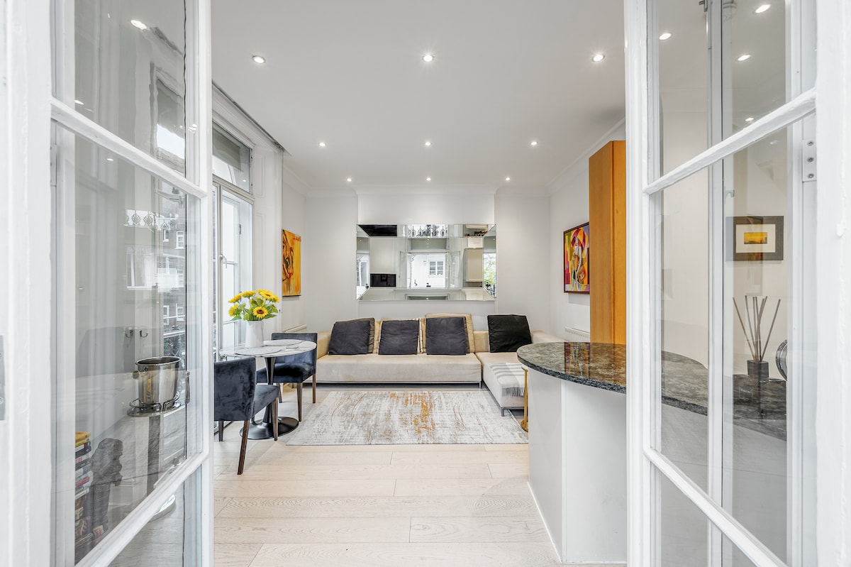 A bright living area features a comfortable sectional sofa and a small dining table. Light streams in from large windows, highlighting the neutral color scheme and modern decor. Artwork is displayed on the walls, adding visual interest to the space.