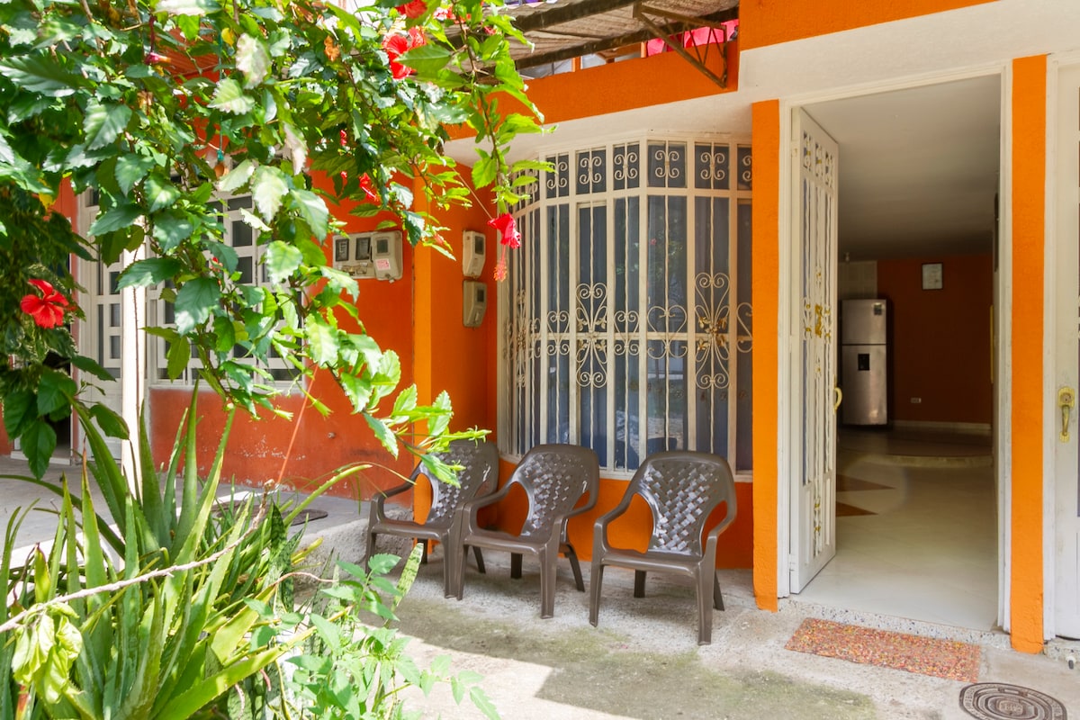 An inviting entrance features three dark chairs arranged on a concrete patio, surrounded by vibrant green plants and blooming flowers. A decorative gate with intricate detailing opens to a brightly colored interior, characterized by a warm orange hue and spacious layout.