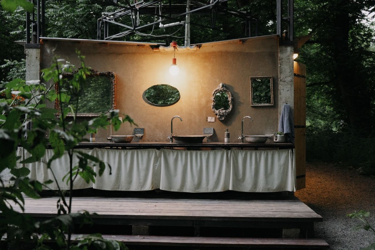 An outdoor wash area is presented with rustic charm, featuring elegant mirrors above three sinks. Soft, flowing fabric covers the base of the counter, complemented by a warm overhead light. The natural surroundings are subtly integrated, enhancing the serene atmosphere.