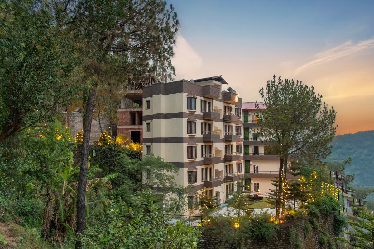 The exterior of the building showcases a multi-story structure nestled among trees, with balconies and large windows. Soft outdoor lighting enhances the greenery around the property, while a scenic view of the hills is visible in the background, illuminated by the colors of sunset.