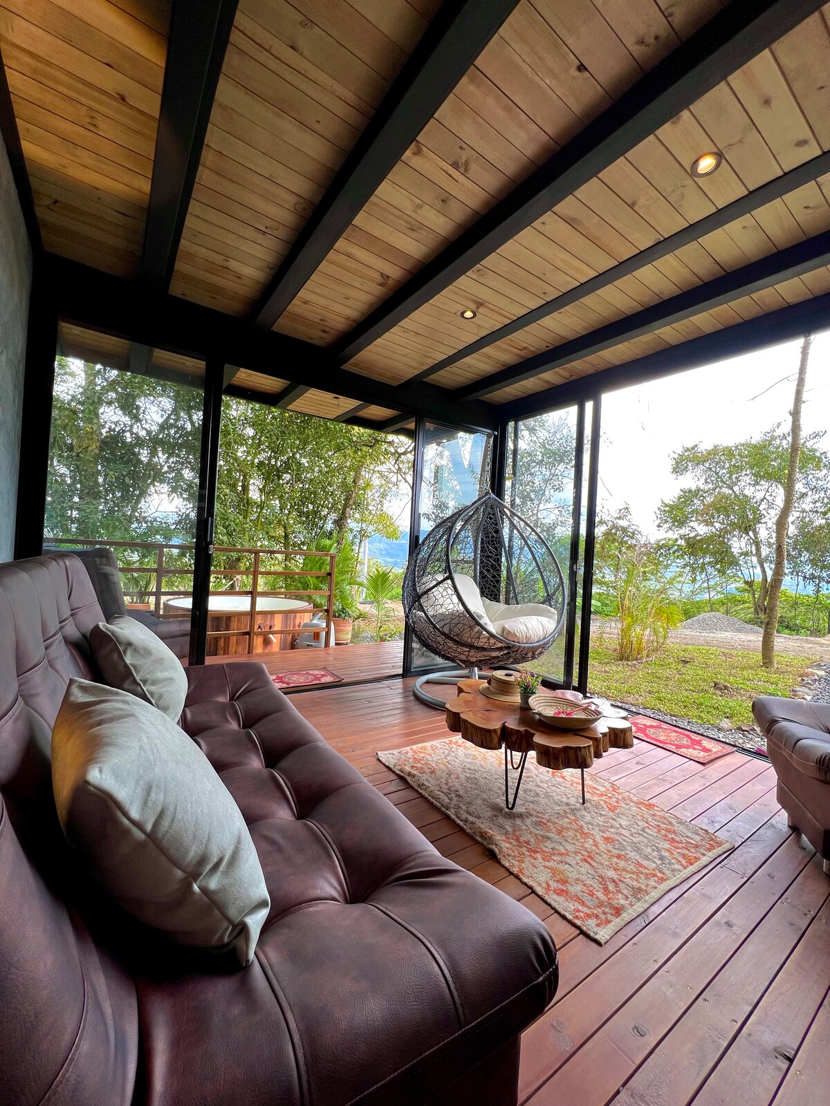 A cozy living area features comfortable seating with plush cushions and a unique hanging chair. Large windows allow natural light to illuminate the space, while views of lush greenery are visible just outside. A decorative rug adds warmth, complemented by a wooden coffee table.