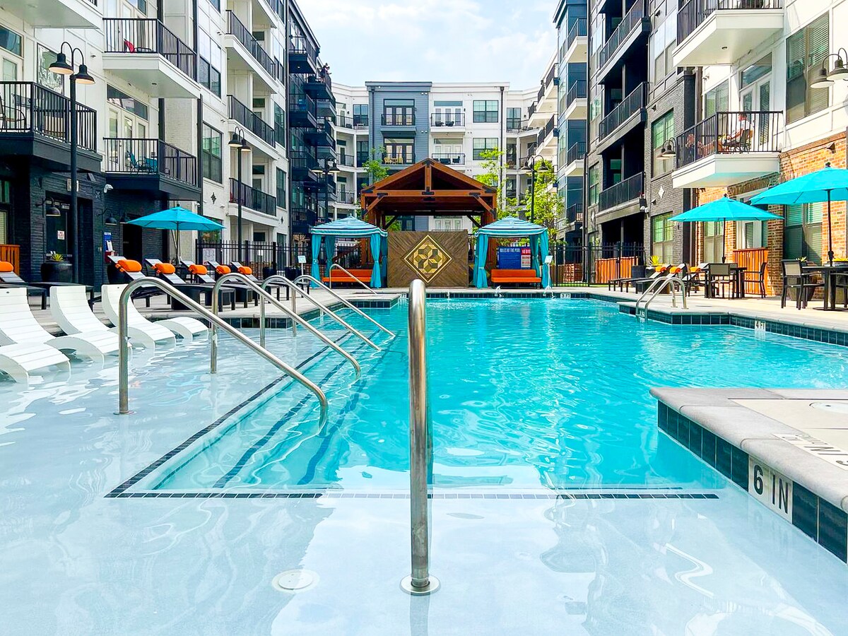A large outdoor swimming pool is surrounded by lounge chairs and shaded areas, providing a refreshing atmosphere. Bright umbrellas offer shade over seating arrangements nearby. The pool area is framed by modern apartment buildings, creating a vibrant courtyard setting.