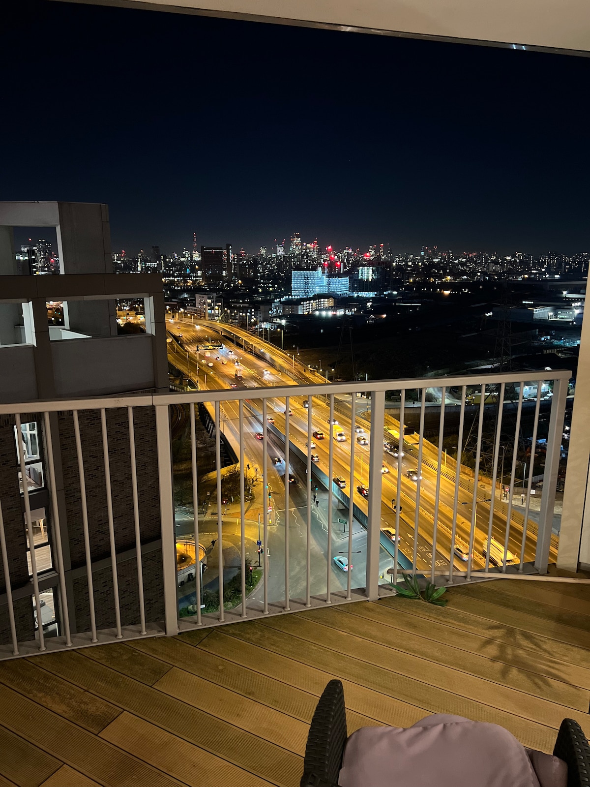 A balcony view displays a vibrant city skyline illuminated at night. The image captures busy roads with vehicle lights creating a dynamic pattern, framed by the railing of the balcony. The wooden flooring adds a touch of warmth to the outdoor space.