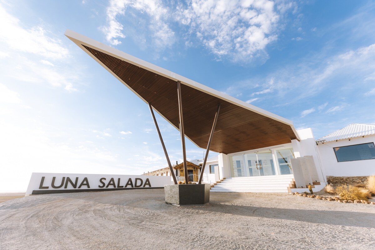 The entrance of the property features a modern architectural design, with a large, slanted wooden roof supported by a central pillar. The name 'LUNA SALADA' is prominently displayed on a stone structure, set against a serene backdrop of the expansive salt flats and blue sky.