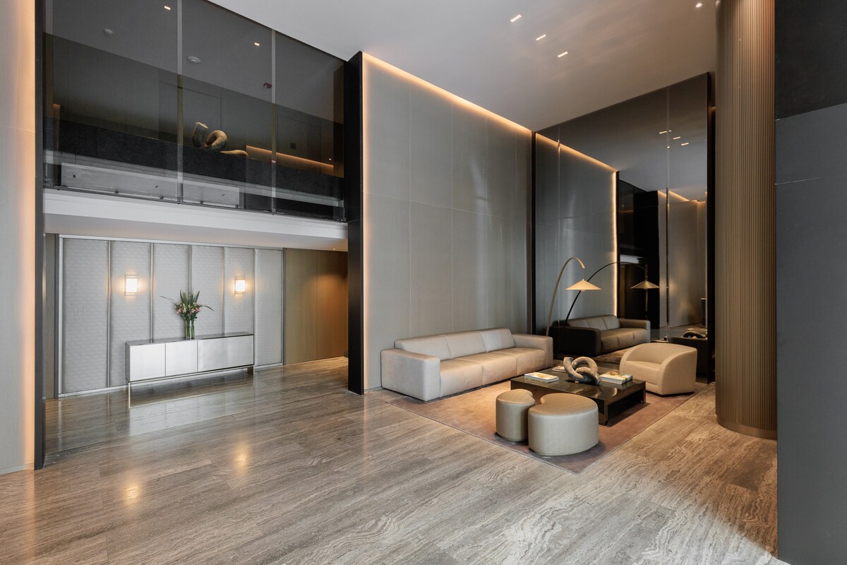 A spacious lobby features polished marble flooring and modern furnishings. A sleek, light-toned sectional sofa is positioned beside a unique coffee table. Ambient lighting enhances the elegant atmosphere, with decorative plants adding a touch of greenery to the contemporary space.