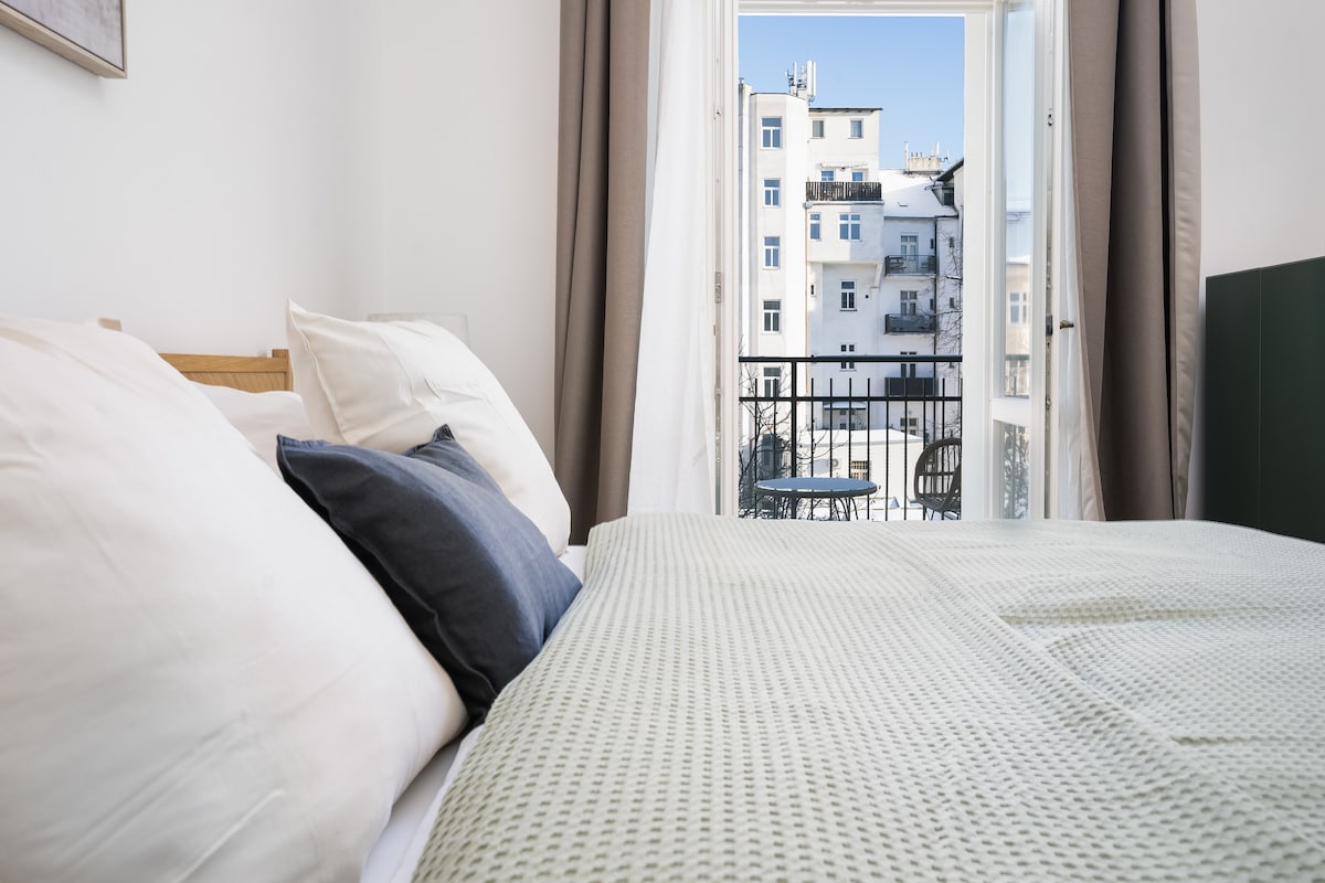 A spacious bedroom features a large bed with soft bedding. Natural light fills the room through a set of double windows that open to a private balcony. The view includes neighboring buildings and outdoor seating arranged on the balcony.