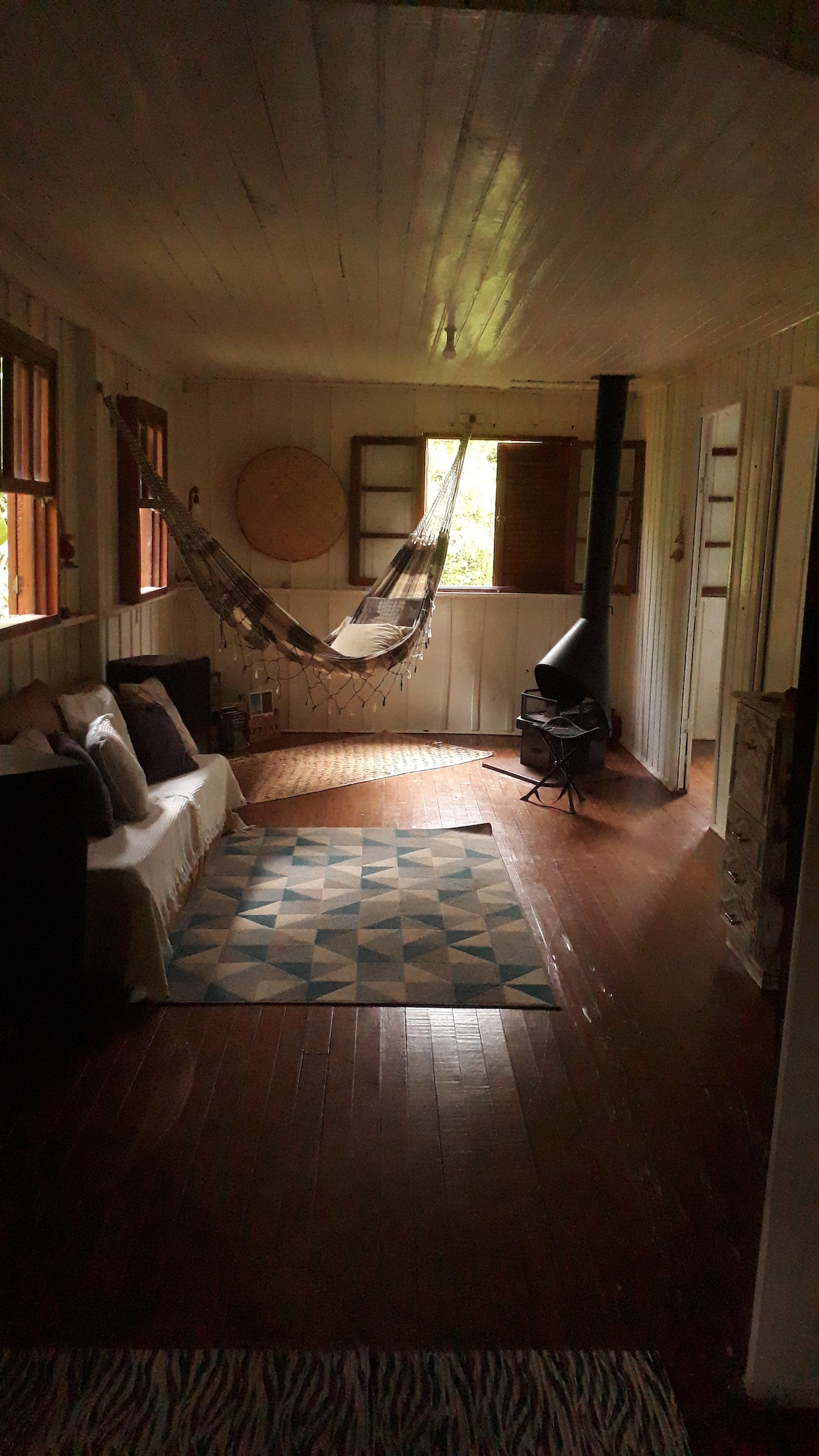 A welcoming interior space features a comfortable seating area with light-colored cushions. A hammock is suspended nearby, and a wood-burning stove is positioned against one wall. Natural light filters through multiple windows, illuminating the wooden flooring and woven area rug.