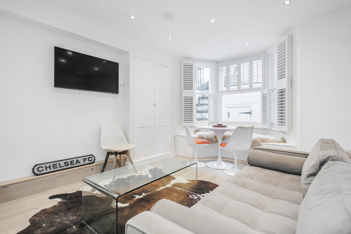 A modern living area is showcased with a comfortable sectional sofa and a glass coffee table. A flat-screen TV is mounted on the wall. The bay window features seating and is fitted with plantation shutters, allowing for natural light to softly illuminate the space.