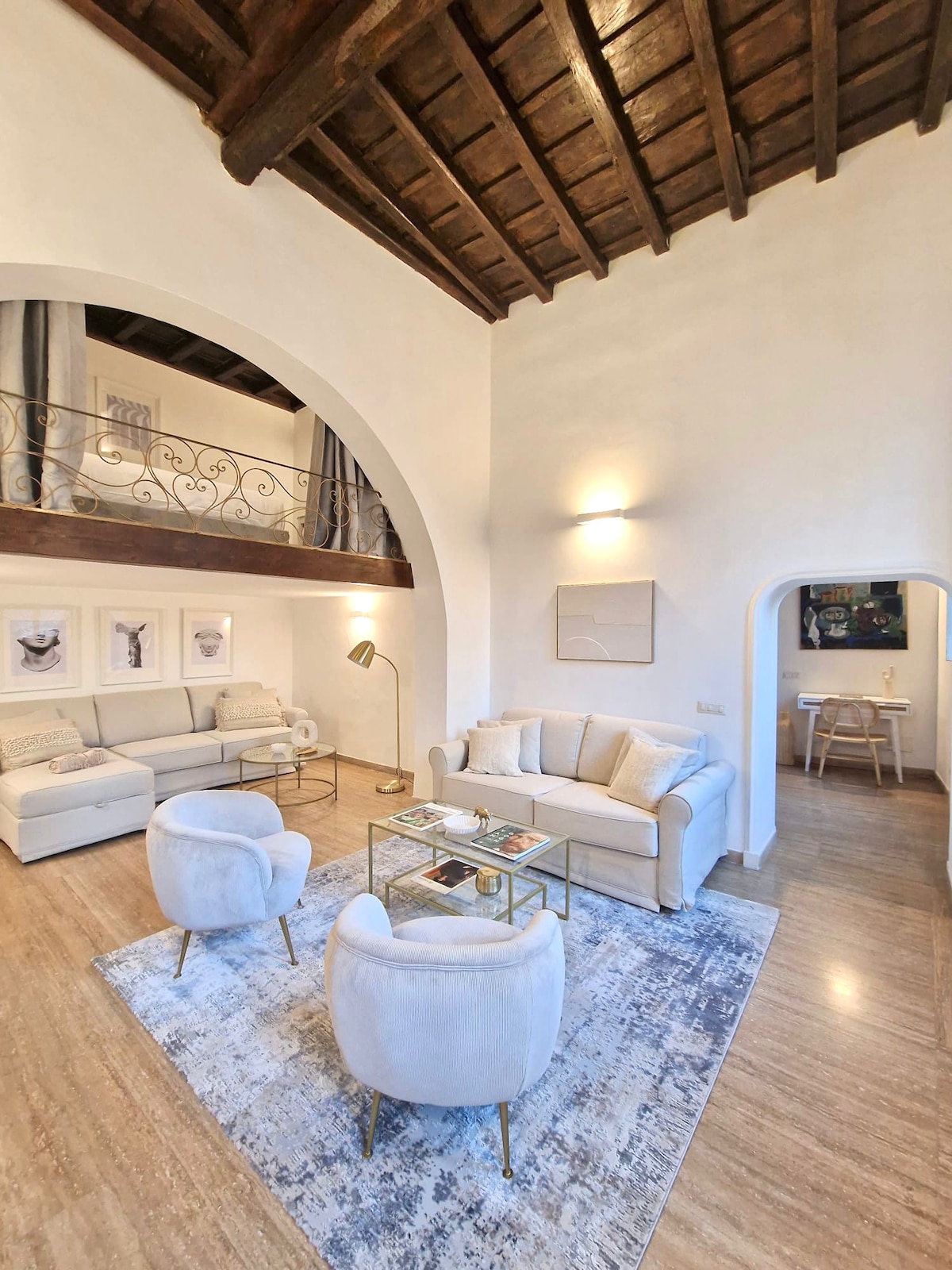 The living area showcases a light-toned sofa and two plush armchairs arranged around a glass coffee table on a soft area rug. Framed artwork is displayed on the adjacent wall, and a high ceiling features wooden beams, adding an airy feel to the space.