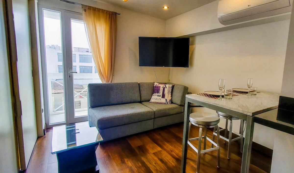 A modern living area is showcased with a comfortable gray sofa and a wall-mounted television. A small dining table with white stools complements the space, while large windows allow natural light to fill the room. Light-colored curtains softly frame the windows.