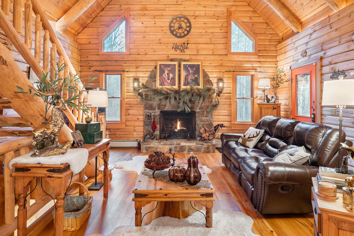 A spacious living area is highlighted by wooden walls and a stone fireplace, which is adorned with greenery. Comfortable leather seating surrounds a wooden coffee table, while decorative items and soft furnishings contribute to the cozy ambiance. Large windows allow natural light to illuminate the room.