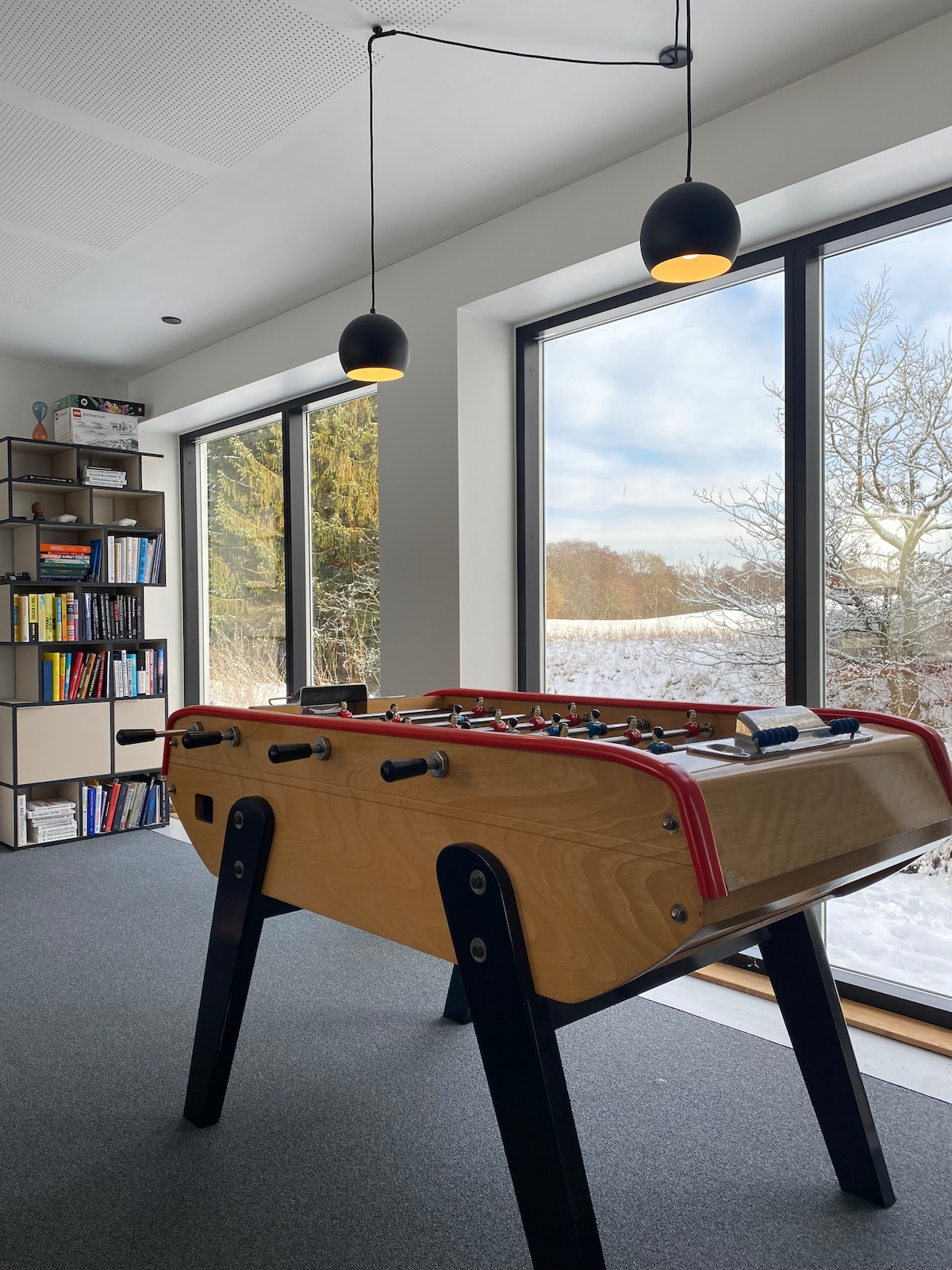 A foosball table equipped with red accents is positioned prominently in the foreground. Behind it, large windows provide natural light and showcase a snowy landscape and trees outside. A bookshelf filled with various books stands to the side, enhancing the leisure space.