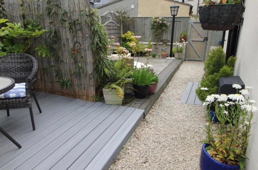 A private garden space is depicted with a combination of decking and pebbled paths. Potted plants and flowers are arranged throughout the area, with a wicker chair placed on the deck, offering a tranquil setting. A wooden gate and fence provide privacy.