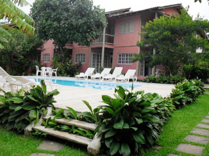 A tranquil pool area is surrounded by lush greenery, featuring multiple sun loungers arranged neatly around the water. The pink facade of the two-story structure is partially visible in the background, framed by trees and plants, creating a serene outdoor environment.