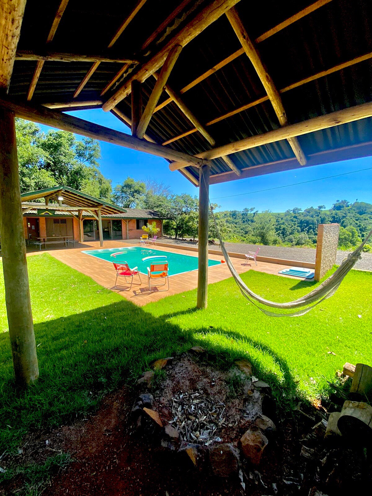 A shaded area features a hammock and two chairs set around a fire pit, with a private pool visible in the background. Lush greenery surrounds the space, providing a serene atmosphere. A covered pavilion is seen to the left, complementing the outdoor setting.