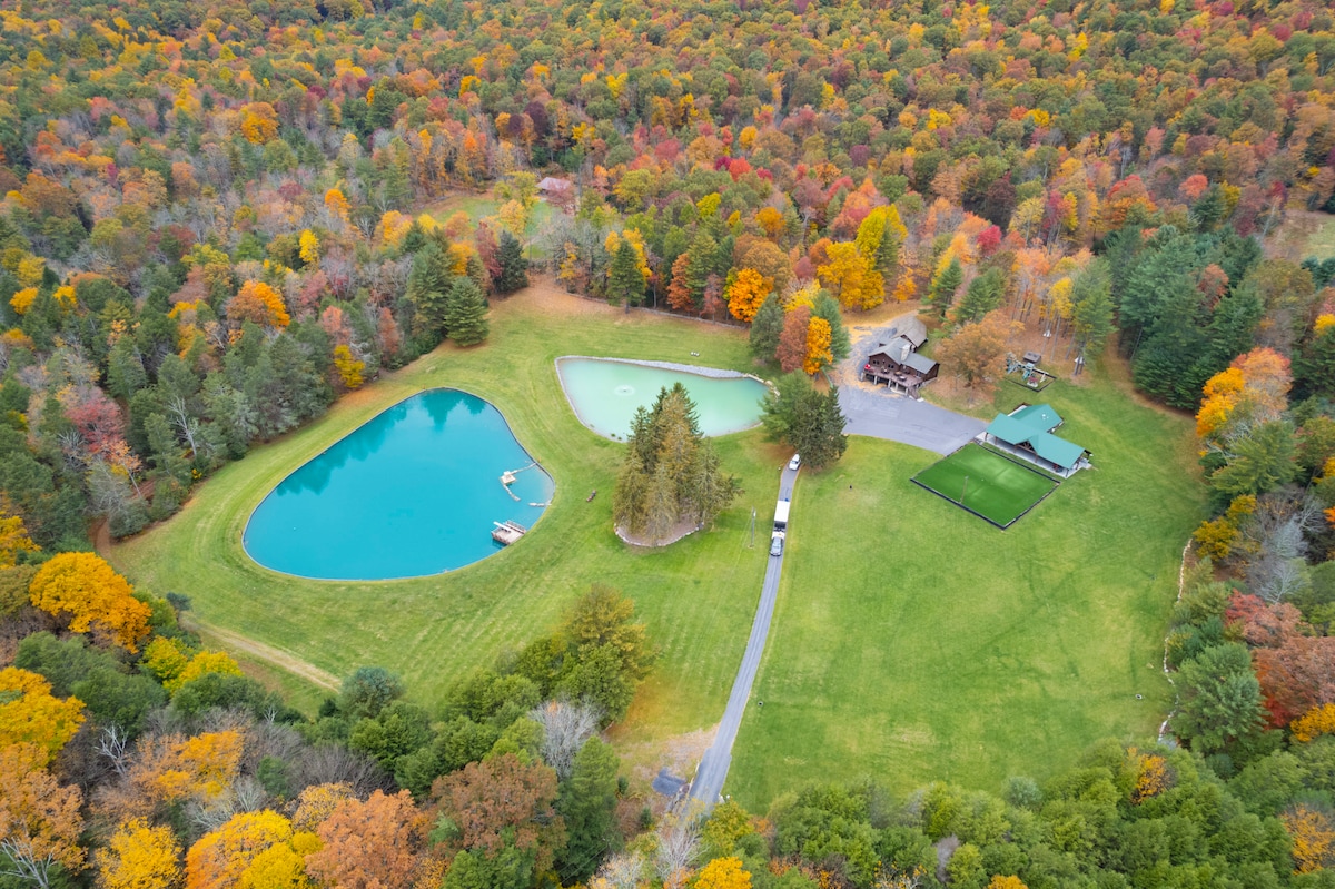 An aerial view captures the expansive property surrounded by vibrant autumn foliage. A large pond is visible, along with a spacious lawn featuring a pavilion, outdoor kitchen, and various recreational areas. The serene landscape promotes a sense of tranquility and connection to nature.