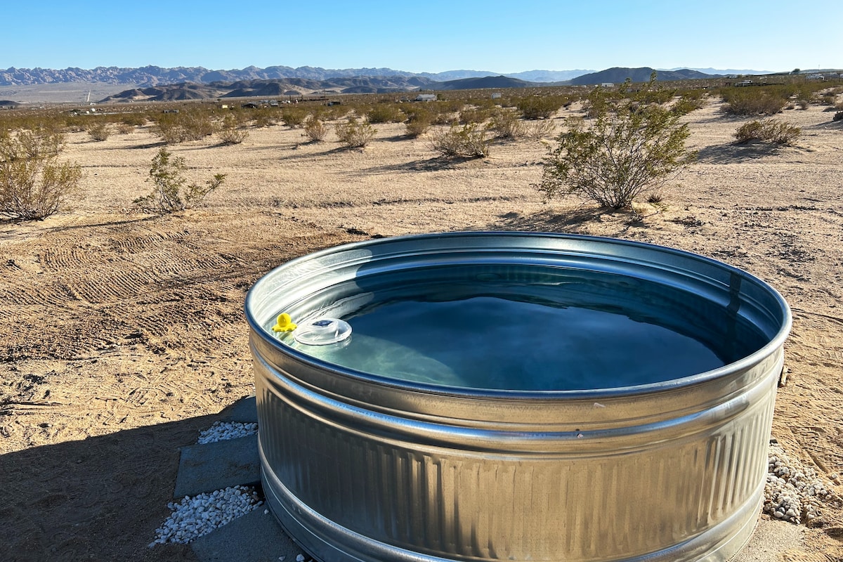 A circular cowboy pool is visible, reflecting the clear skies and surrounding desert landscape. The pool's metallic edges contrast with the dry terrain, and distant mountains can be seen in the background, emphasizing the peaceful outdoor setting.