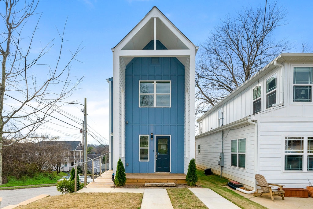 A contemporary two-story home is showcased with a blue gabled exterior. The entrance is framed by two small trees and a landscaped area with wooden steps leading to the door. The surrounding area features a clear sky and well-maintained lawns.
