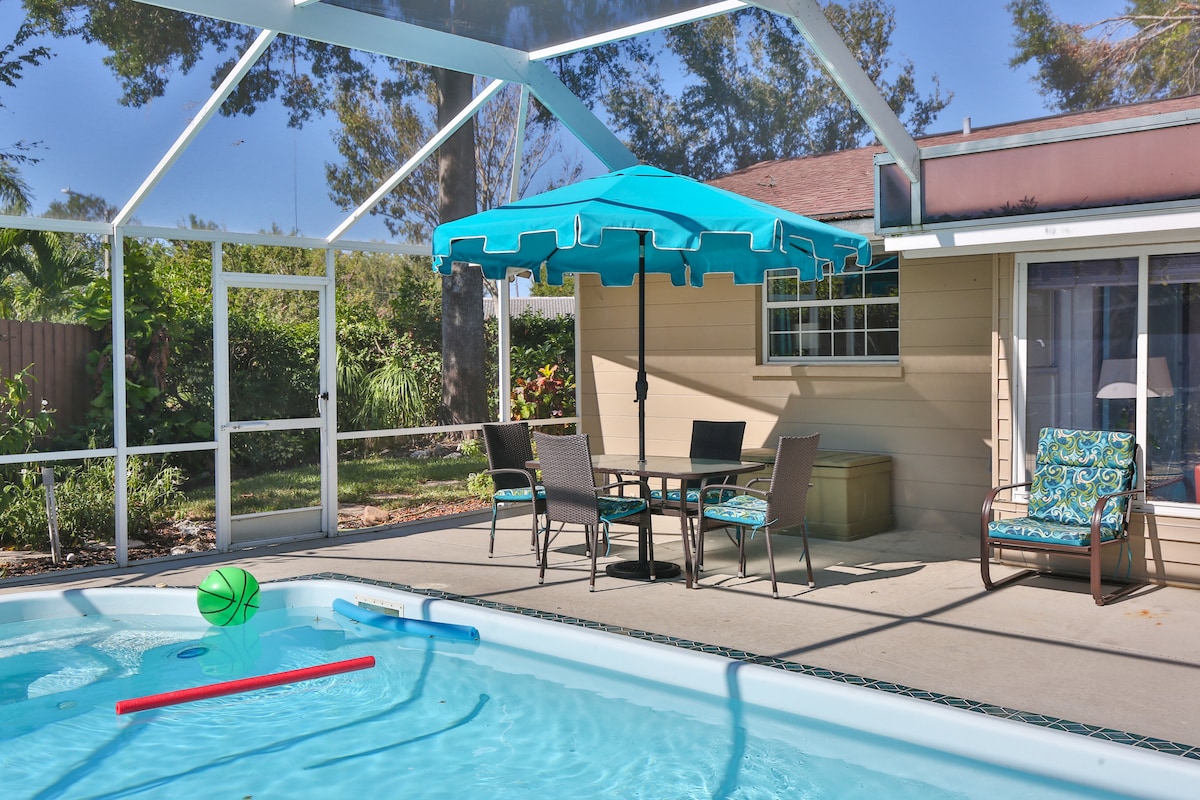 An outdoor space is featured, showcasing a private pool surrounded by a screened enclosure. A patio table with chairs is positioned near a large umbrella, providing shade. Colorful pool toys float on the water, and lush greenery creates a tranquil backdrop.