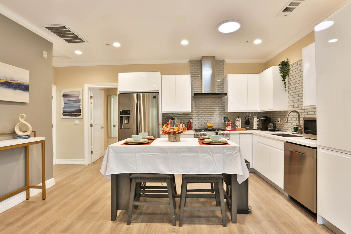 The bright kitchen showcases white cabinetry with modern stainless-steel appliances. A large table is set with placemats and dinnerware, while a stylish backsplash adds texture to the space. Large windows allow natural light to highlight the warm wood flooring.