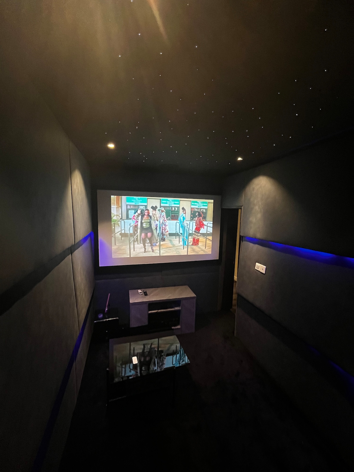A cozy cinema room features a large projector screen displaying a film. Soft lighting from recessed spots and a starry ceiling create a relaxing ambiance. A low coffee table sits in front of a plush seating area, enhancing comfort during movie nights.