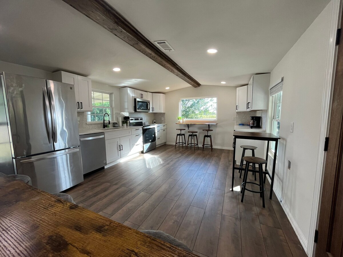 A modern kitchen features stainless steel appliances, including a refrigerator and oven. Natural light filters through windows, illuminating the space. A wooden beam runs across the ceiling, complementing the light-colored cabinetry. Two tall stools are positioned at a compact dining area.