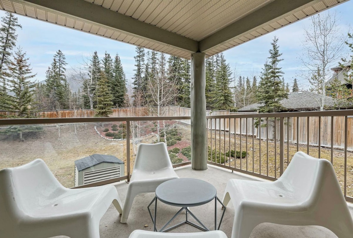 A private balcony features four white chairs arranged around a circular table. The surrounding area includes a grassy yard with trees and shrubs, creating a serene outdoor space for relaxation.