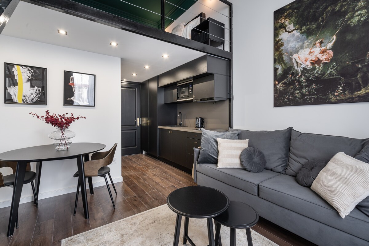An inviting living space features a cozy sofa, two small black tables, and a round dining table with four chairs. The kitchenette, equipped with modern appliances, is visible in the background. Artwork is displayed on the walls, enhancing the contemporary decor.