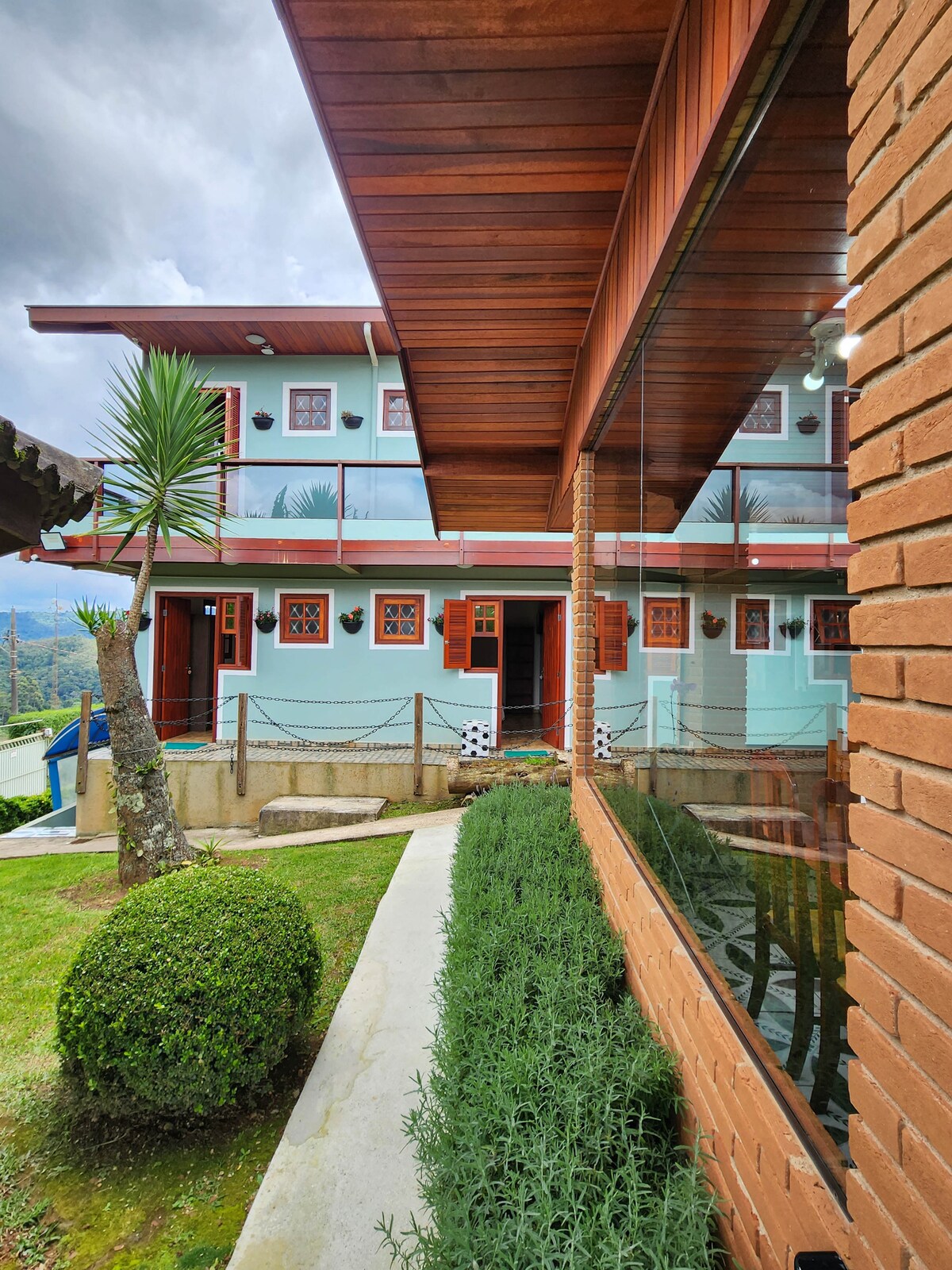 The exterior of the pousada features a light blue façade adorned with wooden window shutters. A well-maintained garden with neatly trimmed hedges lines the pathway, and a wooden overhang provides a shaded area. Scenic views of the surrounding landscape are visible through large glass panels.