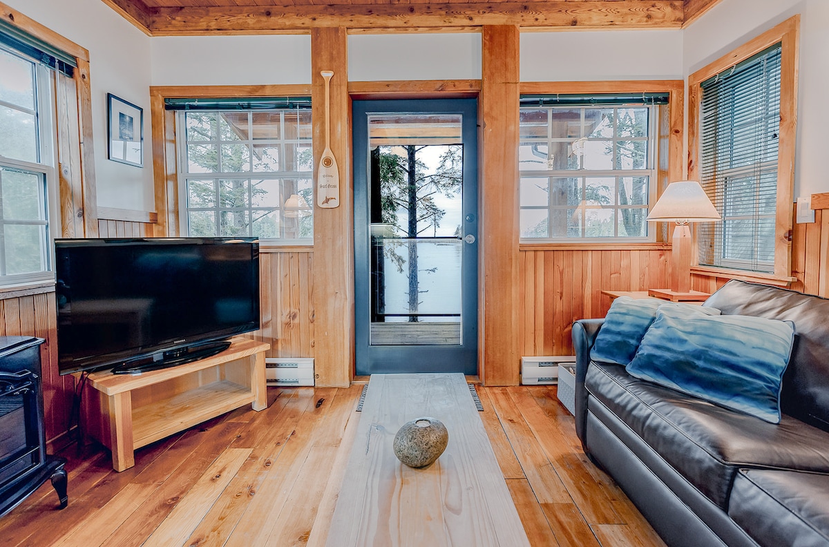 The living area features large windows inviting natural light and offering views of the surrounding landscape. A comfortable leather sofa is adorned with decorative pillows, while a wooden coffee table sits centrally. A television is on a wooden stand across from the seating area.