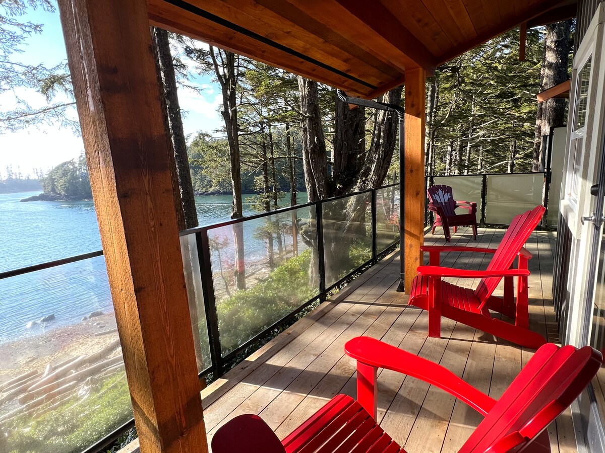 A spacious deck framed by wooden beams overlooks the tranquil waters and surrounding trees. Two red Adirondack chairs provide a comfortable seating area, inviting relaxation amidst the natural scenery.