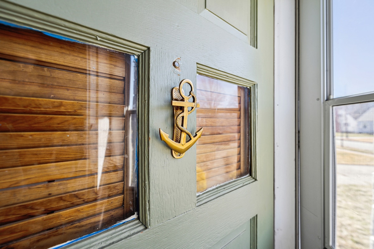 A green front door is adorned with a decorative anchor motif. Light filters through two small windows, highlighting the wood paneling behind. The exterior setting features a view of a residential neighborhood, creating an inviting entrance.