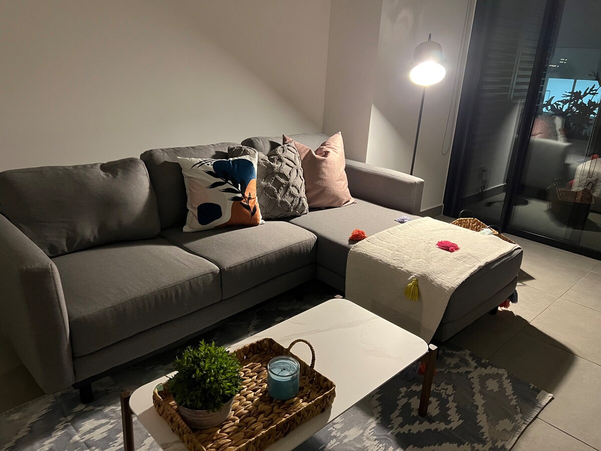 A modern living area is presented, featuring a spacious gray sectional sofa adorned with colorful cushions. A low coffee table with a decorative tray sits in front of the sofa, complemented by a soft throw. Natural light filters through large glass doors.
