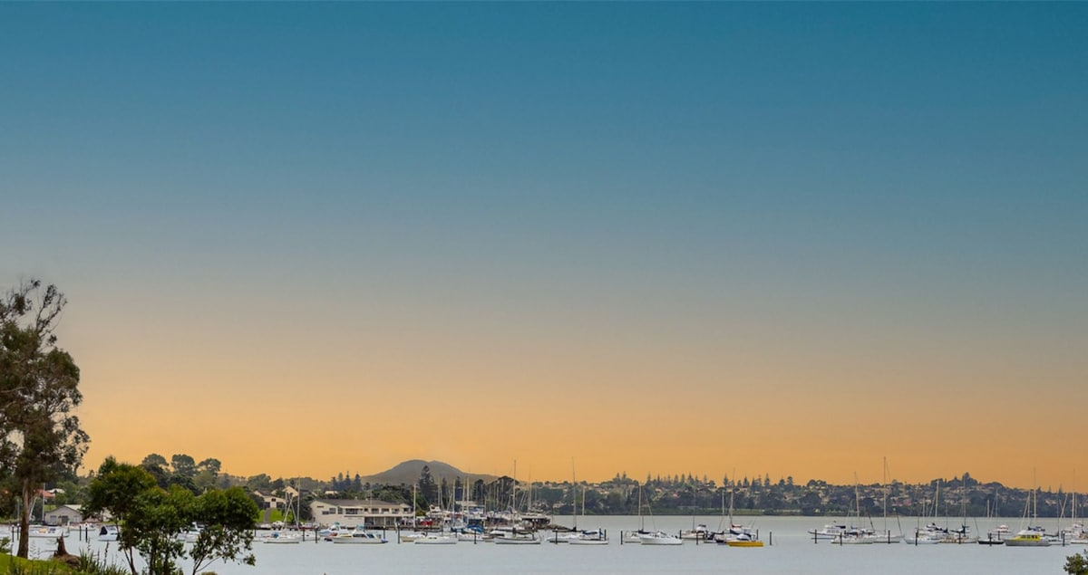 A serene waterfront scene captures the tranquil waters of Tamaki Bay, dotted with boats at anchor. The horizon displays a gradient of soft pastel colors as the sun sets, framed by distant hills and lush greenery along the shore.