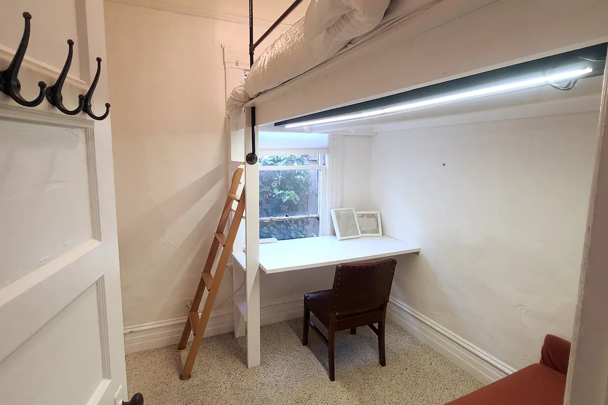 A cozy room is highlighted by a lofted bed above a functional workspace. Below, a desk is paired with a chair, and soft lighting illuminates the area. A decorative ladder leads to the loft, while a sofa sits near a window that offers natural light.