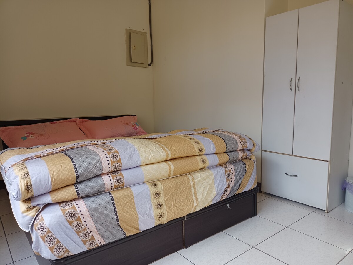 A bedroom features a spacious bed with layered comforters in muted tones. A simple white wardrobe is positioned nearby, offering additional storage. Neutral-colored walls and tiled flooring create a clean and open environment.