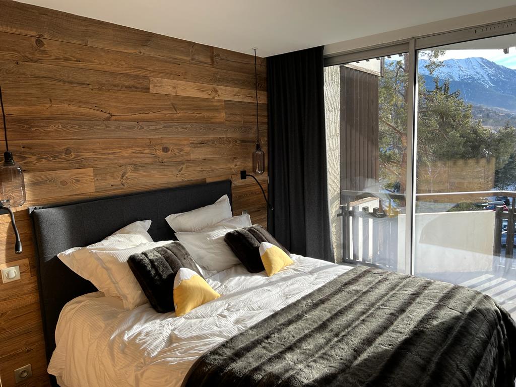 A cozy bedroom features a spacious bed adorned with decorative pillows. Large glass doors open to a balcony, allowing natural light to fill the room. Wooden paneling adds warmth to the space, complemented by dark curtains that frame the view of the mountains outside.