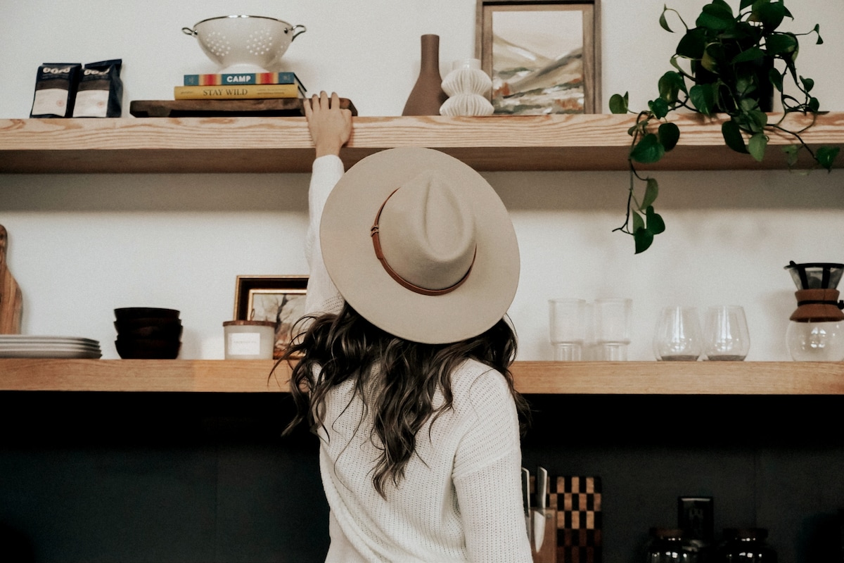 A neatly organized open shelf showcases a variety of items, including a colander, a small vase, and books. A person reaching for an item is depicted, wearing a neutral colored hat. The shelf adds warmth to the kitchen area, complemented by natural light.