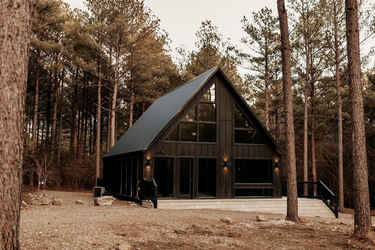 An A-frame cabin is surrounded by tall pine trees, featuring a dark exterior and large glass windows that invite natural light. A spacious deck extends from the cabin, providing a tranquil outdoor space. The natural landscape enhances the secluded atmosphere.