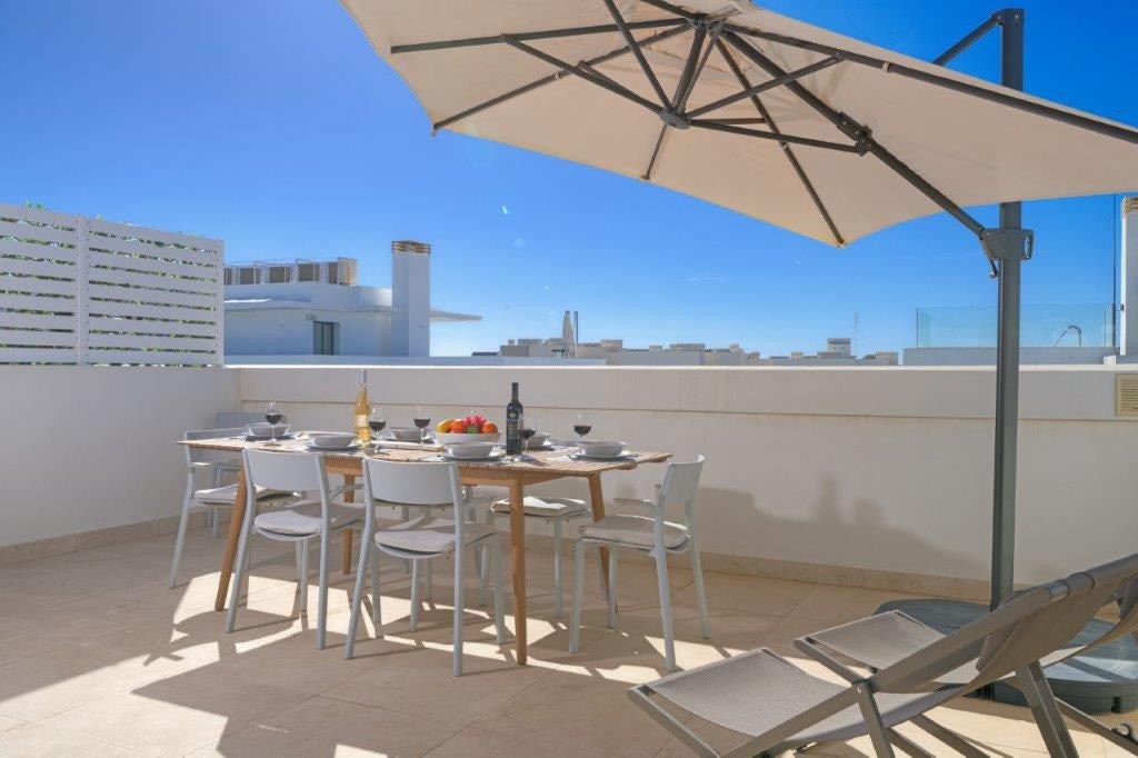 A spacious terrace is displayed with a wooden dining table set for six, accompanied by an umbrella providing shade. Two lounge chairs are positioned nearby, offering a comfortable spot to relax. The clear blue sky creates a serene backdrop for outdoor dining.