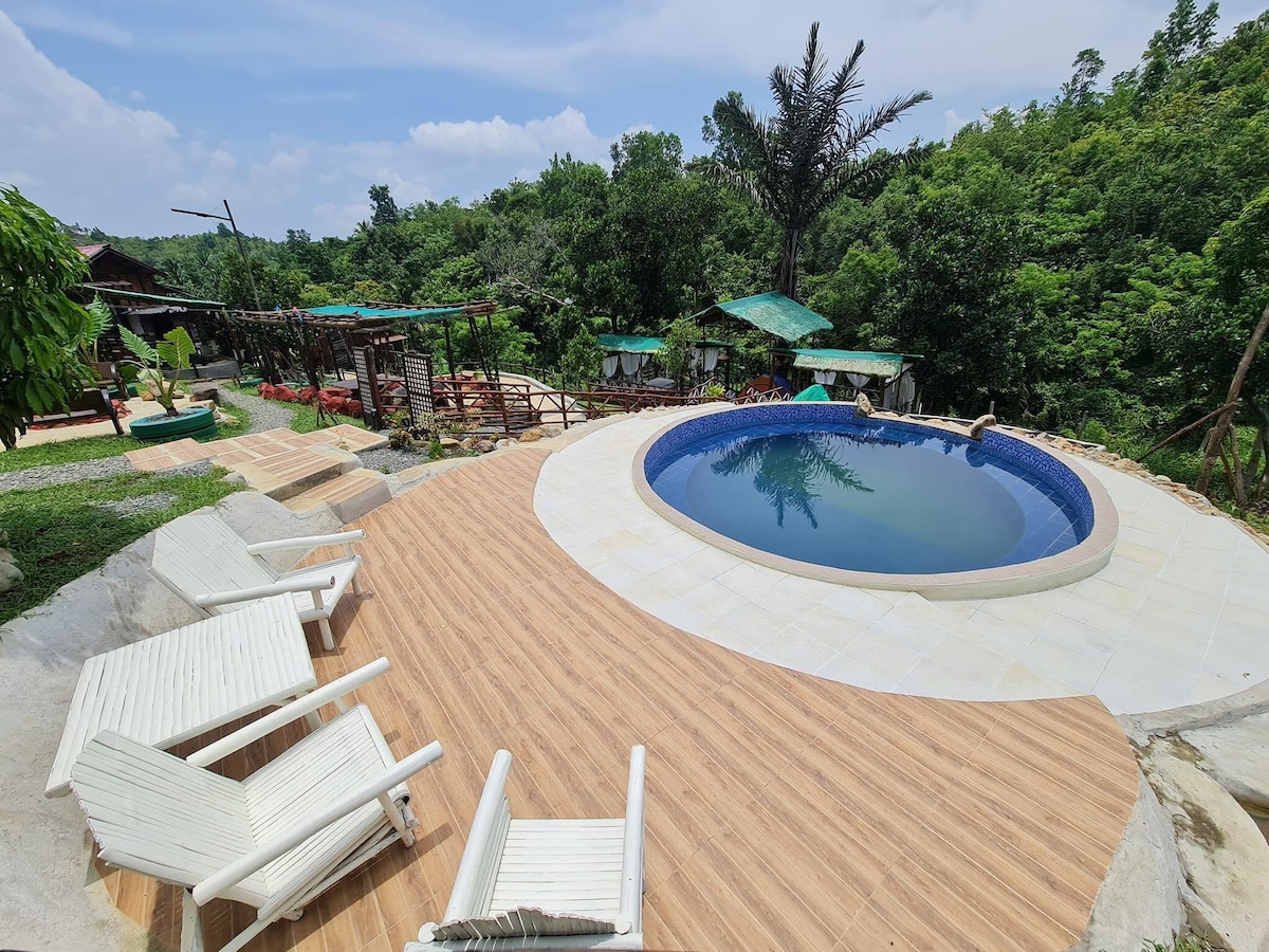 An inviting pool area is set amidst lush greenery, featuring a circular pool surrounded by a wooden deck. Several white lounge chairs are arranged around the pool, offering relaxation spots under the sun. Nearby, trees and canopies provide shade, enhancing the natural environment.